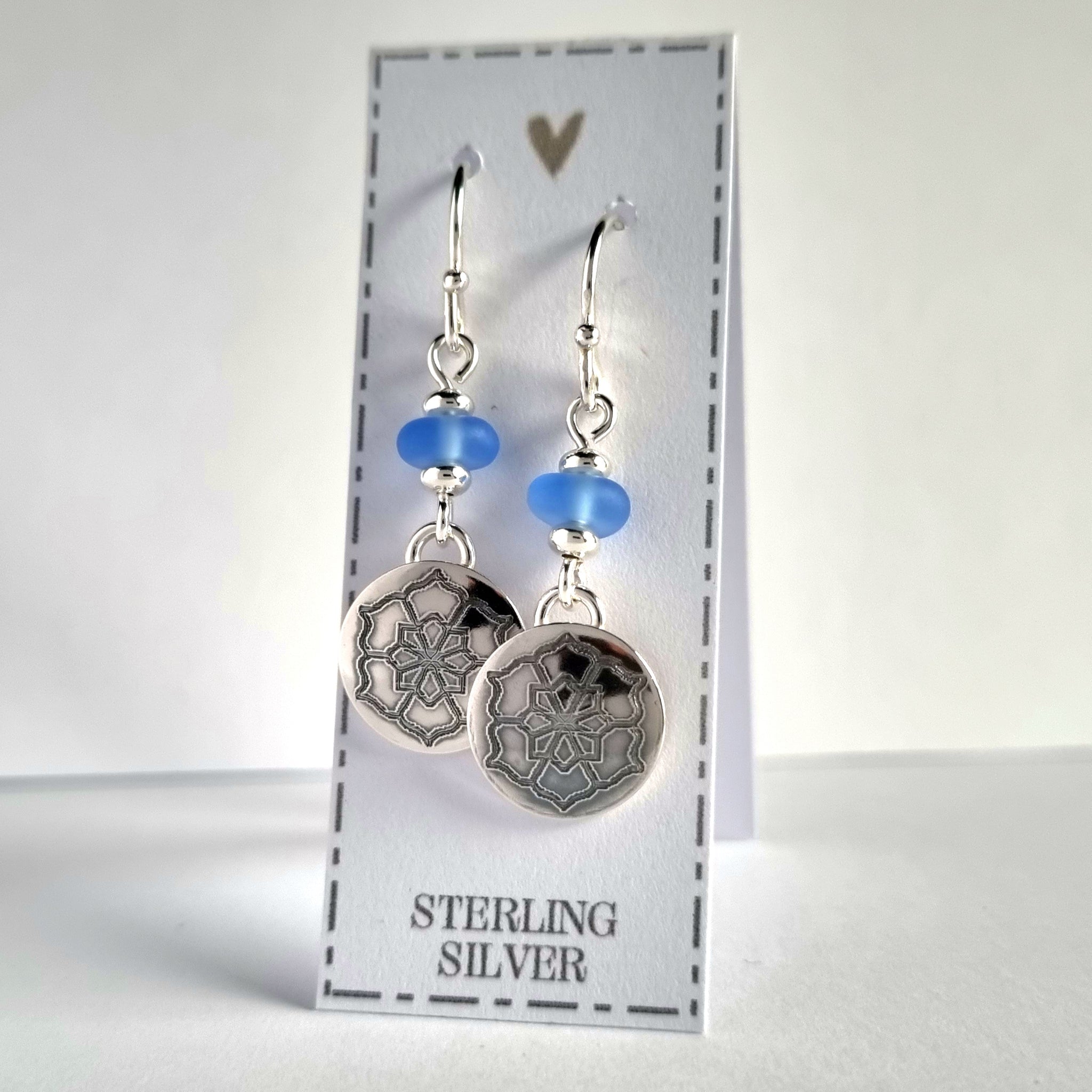 Sterling silver sea glass earrings with engraved flower design. The 12mm engraved silver discs are suspended from the pale blue sea glass beads.