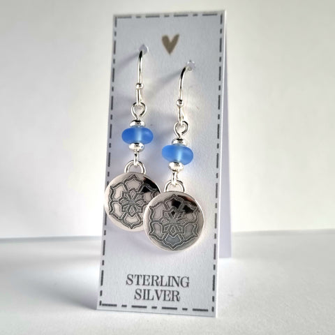 Sterling silver sea glass earrings with engraved flower design. The 12mm engraved silver discs are suspended from the pale blue sea glass beads.