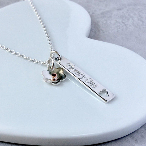 21st birthday necklace, a sterling silver bar pendant engraved with twenty one on the front and the birthday girls name on the back. A silver flower charm is also added to the chain and sits beside the bar.