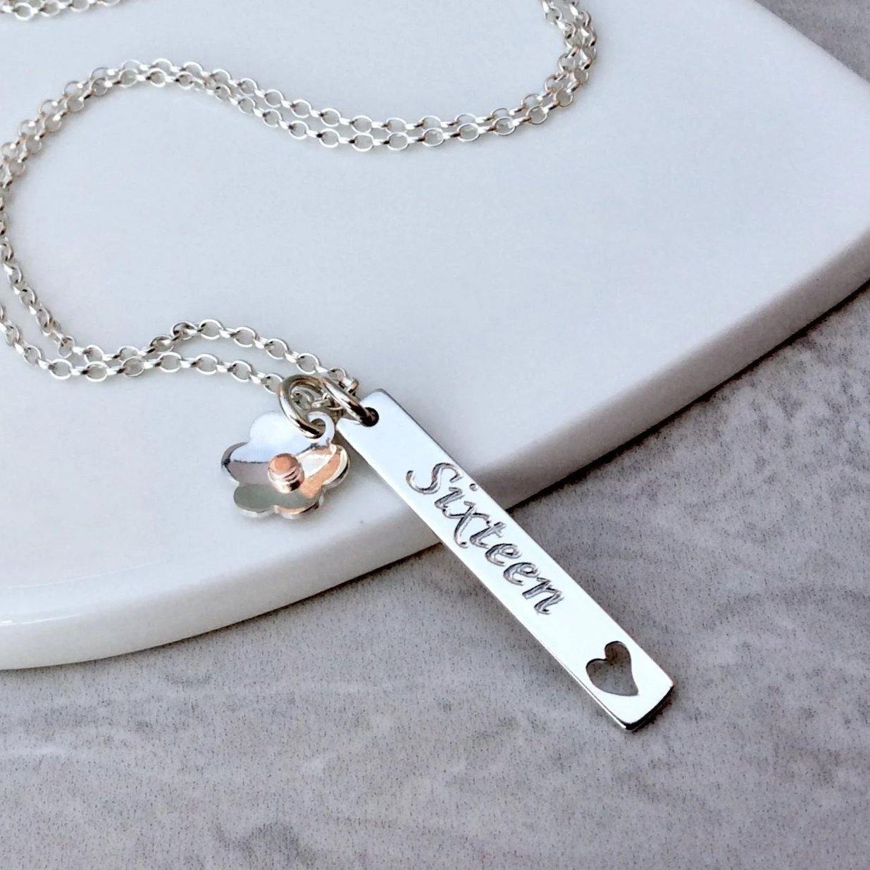 16th birthday necklace, a sterling silver bar pendant engraved with sixteen on the front and the birthday girls name on the back. A silver flower charm is also added to the chain and sits beside the bar.